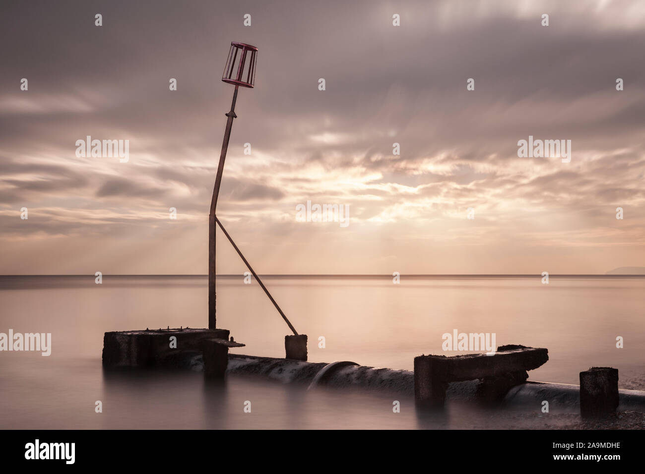 Tuyau d'écoulement et marqueur sur plage avec rayons de soleil crépusculaire à travers les nuages en streaming et longue exposition flou marée montante, Bexhill on Sea, à l'Est de sus Banque D'Images