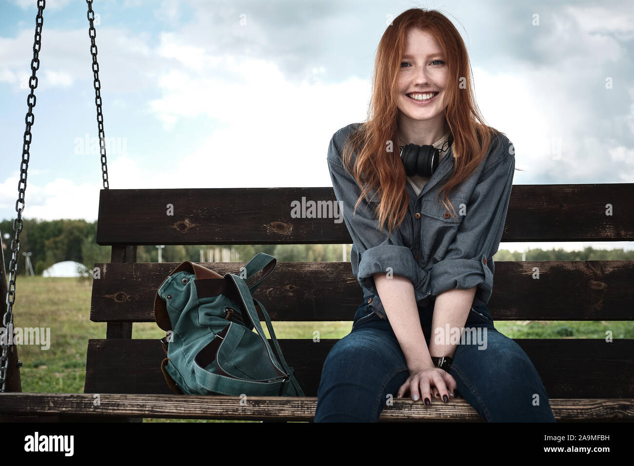 Redhead girl dans une chemise en jean est assis avec un sac à dos sur un banc de bois swing sourit largement à la recherche dans le cadre. Banque D'Images