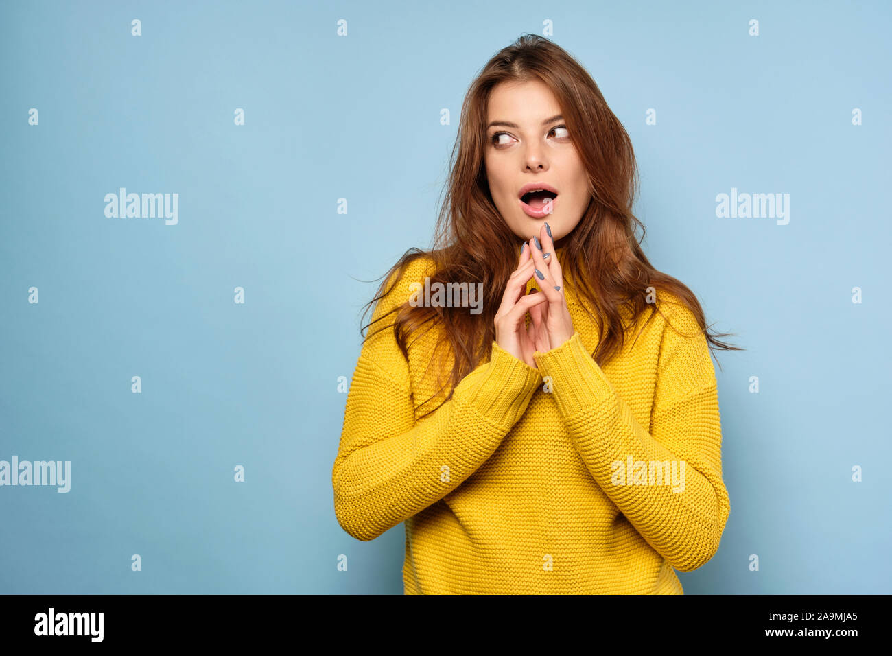 La brunette se dresse sur un fond bleu dans un chandail jaune et regarde ailleurs, ouvrant les yeux et la bouche Banque D'Images