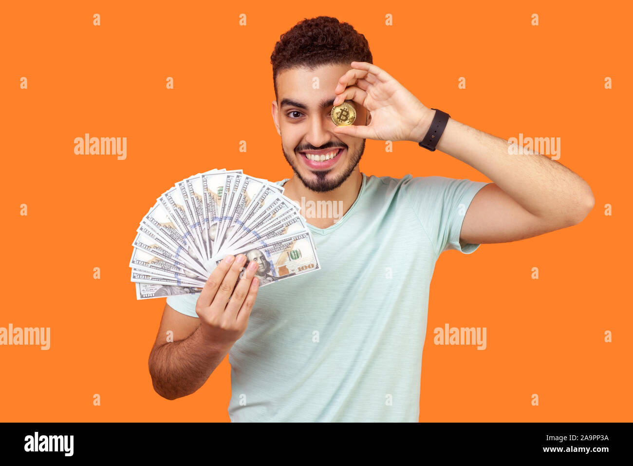 Portrait d'homme riche brunette joyeuse avec barbe en blanc T-shirt holding dollar billets et un œil avec golden ear cryptocurrency, bitcoin Banque D'Images