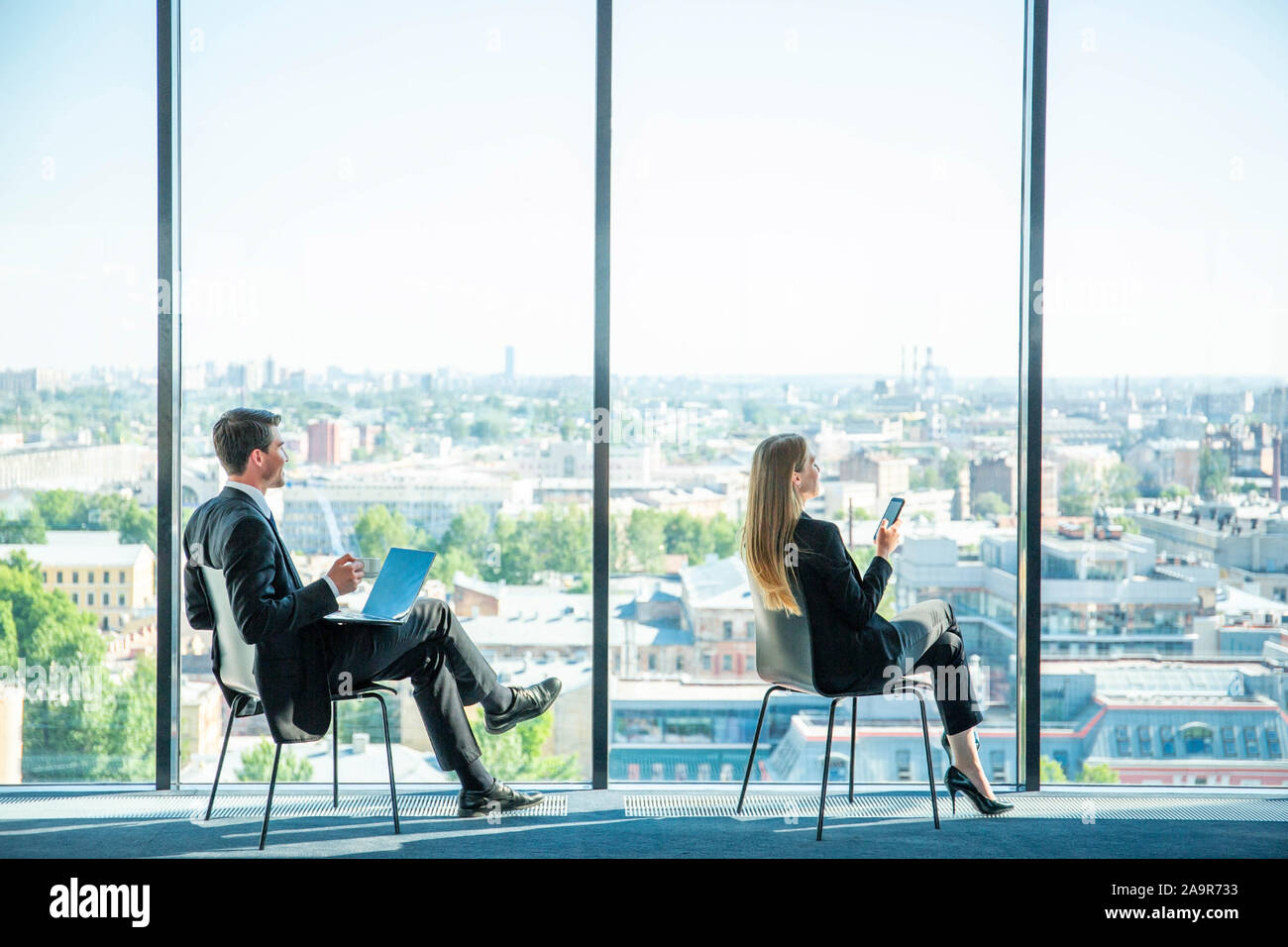 L'homme d'affaires avec ordinateur portable et business woman with smartphone assis près d'une fenêtre de bureau panoramique Banque D'Images