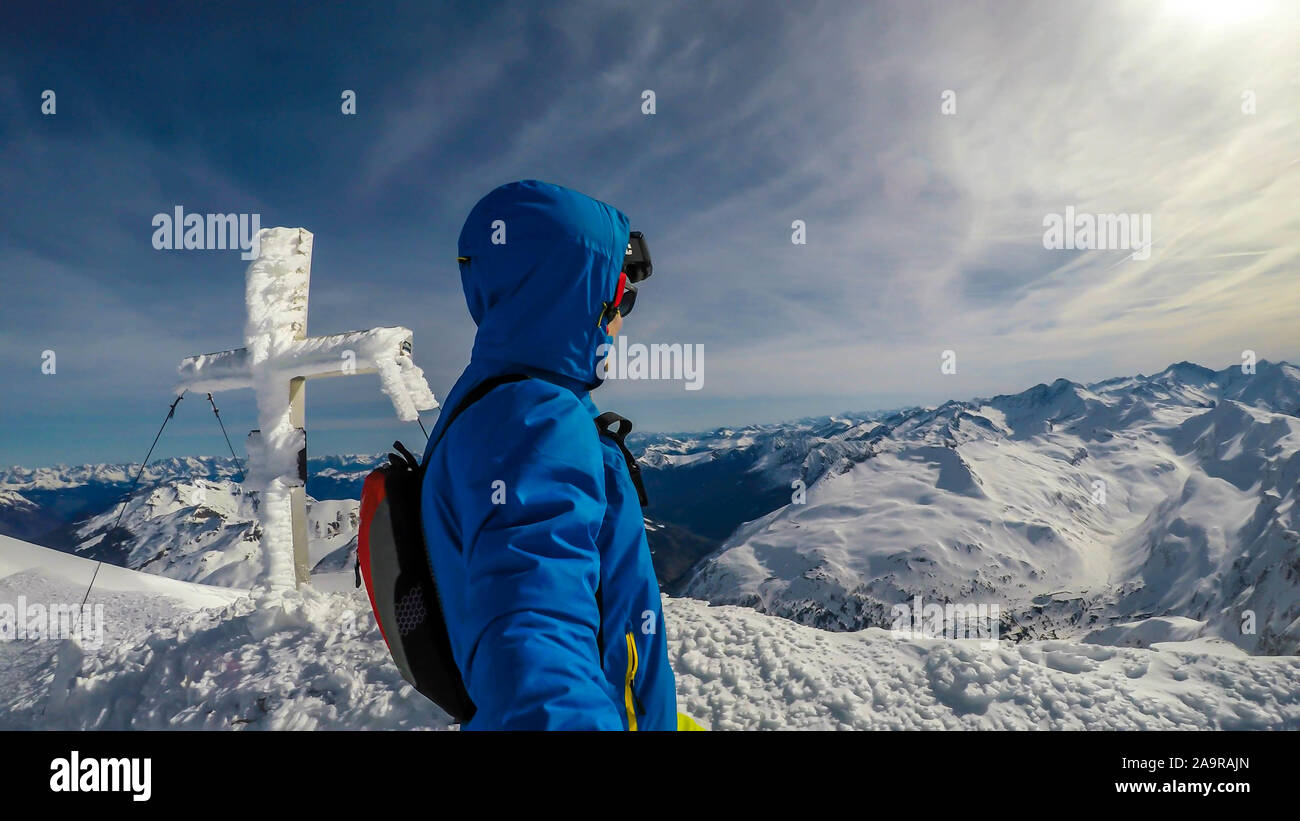 Un homme en tenue de ski debout sur le sommet d une montagne en crete avec une croix couverte de neige derriere lui. La neige sans fin autour de la montagnes caped man