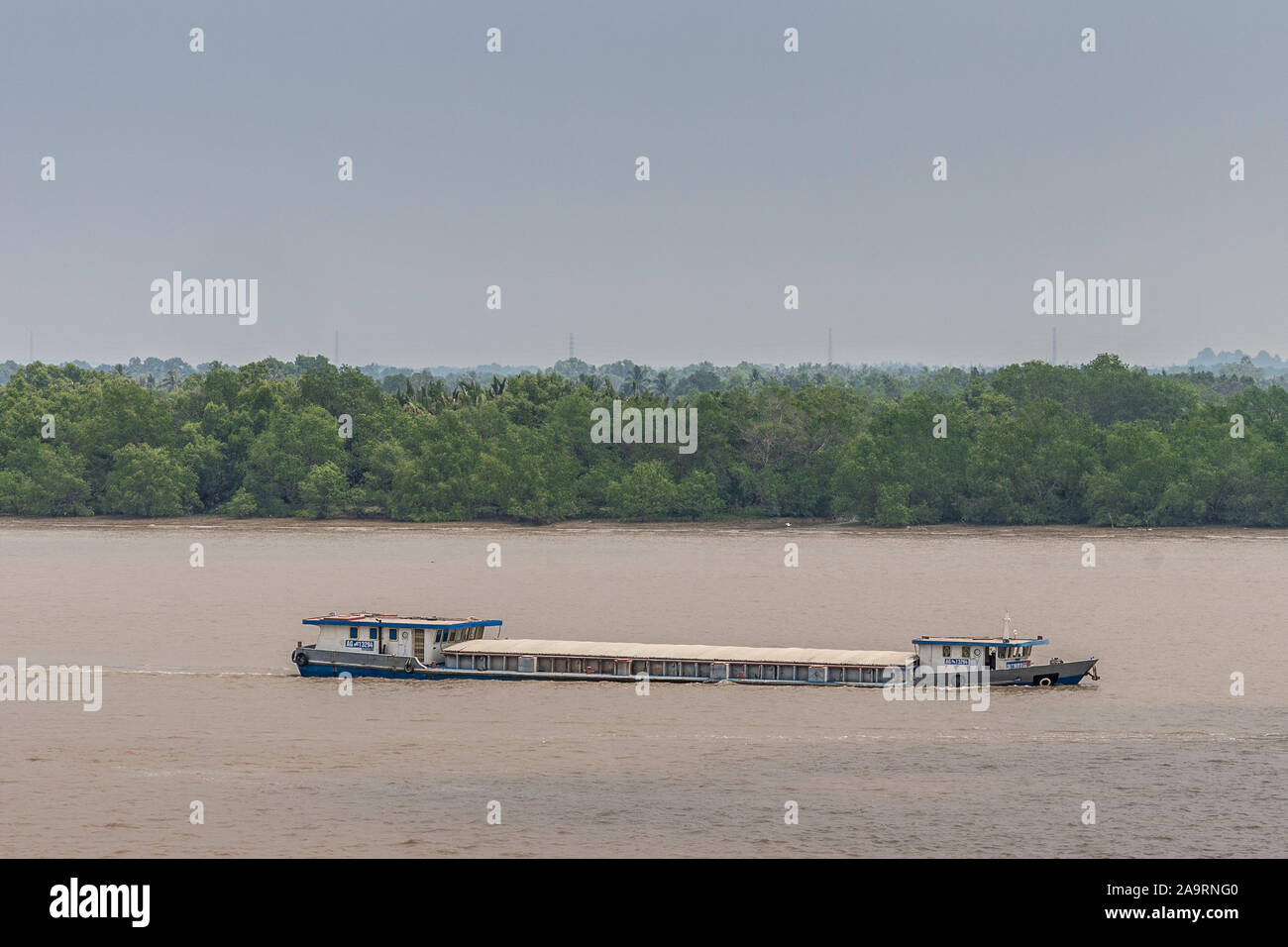 Tau Long River, au Vietnam - 12 mars 2019 : Long hull-fermé river barge voiles sur l'eau brune et se trouve profondément. Ceinture verte se détache du fond de ciel bleu. Banque D'Images