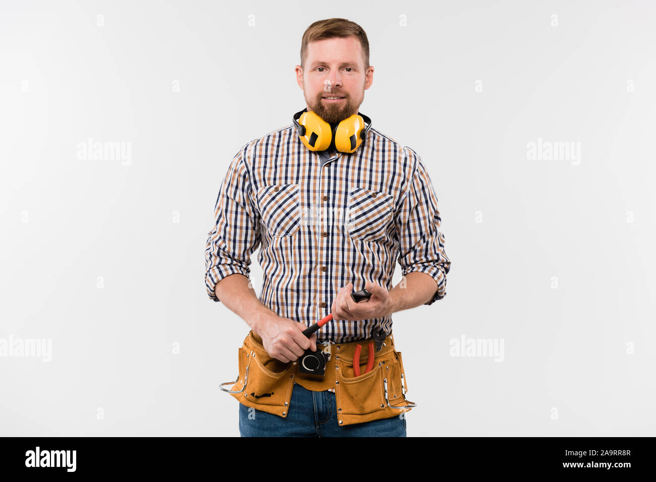 Heureux jeune réparateur barbu de protection avec écouteurs, marteau et ceinture d'outils Banque D'Images