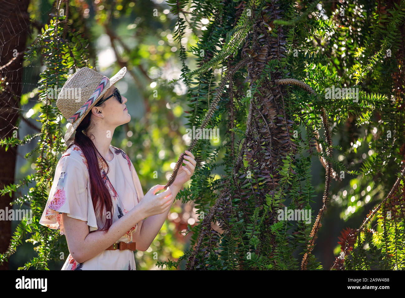 Jolie jeune femme aux longs cheveux rouges portant chapeau de paille, lunettes de soleil et de l'examen d'une robe à fleurs feuilles piquantes Banque D'Images