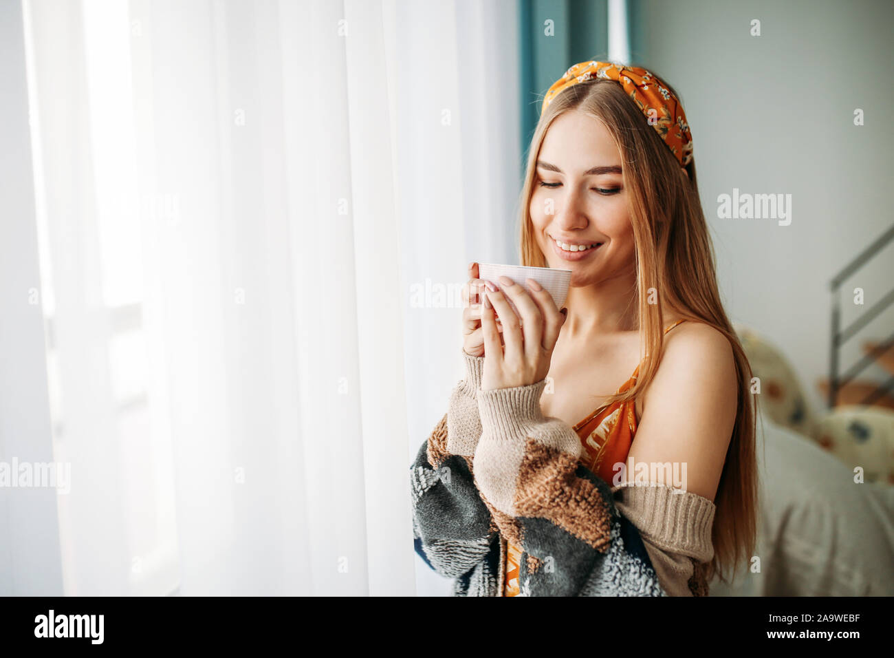 Belle jeune femme cheveux longs juste girl wearing dans la confortable cardigan tricoté avec tasse de thé près de fenêtre à la maison Banque D'Images