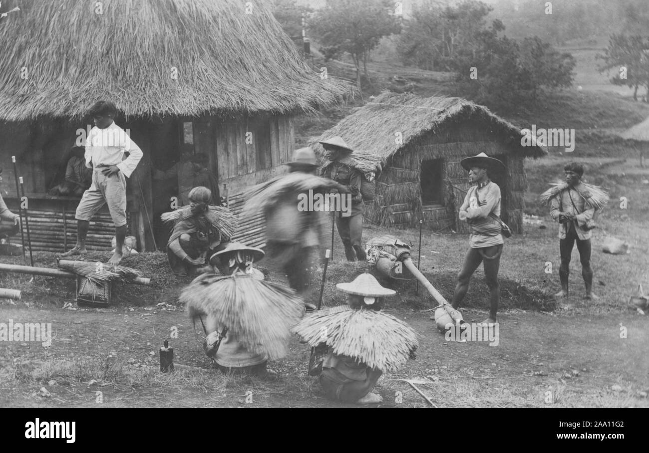 Carte postale monochrome d'un groupe d'hommes, certains portant des capes de pluie en chaume, en face de nipa cabanes, l'île de Luzon, aux Philippines, en 1915. À partir de la Bibliothèque publique de New York. () Banque D'Images