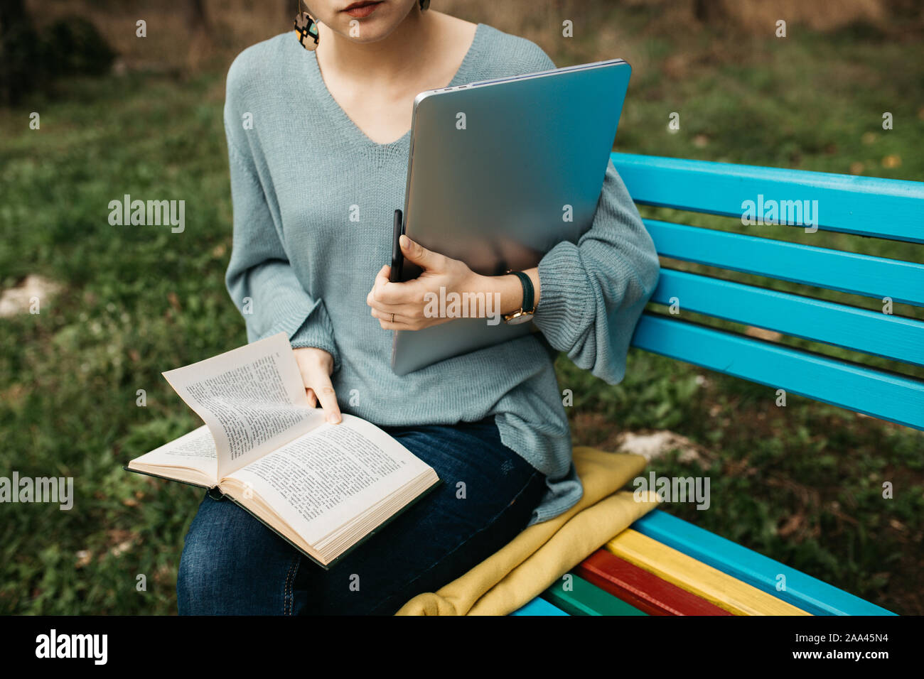 Portrait de femme assise sur le banc en bois coloré en parc. Elle est en train de lire un livre et la tenue d'ordinateur portable et téléphone mobile. Travail freelance concept. Banque D'Images
