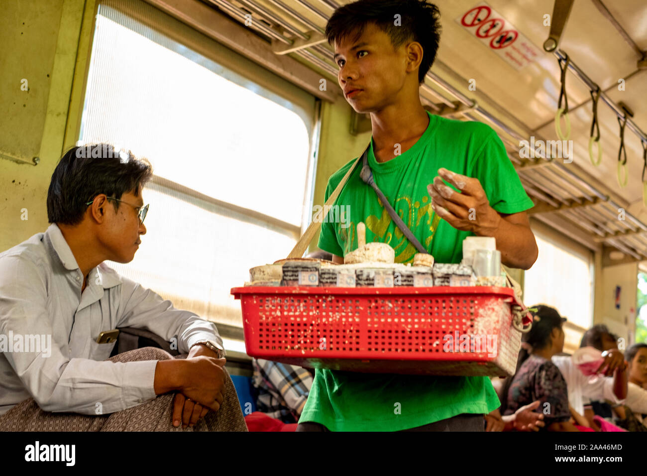 Birman jeune vendeur vend ses marchandises vers le bas-côté d'un train à partir d'un panier en plastique tout en montant le train qui encercle le centre de Yangon, Myanmar (Birmanie) Banque D'Images