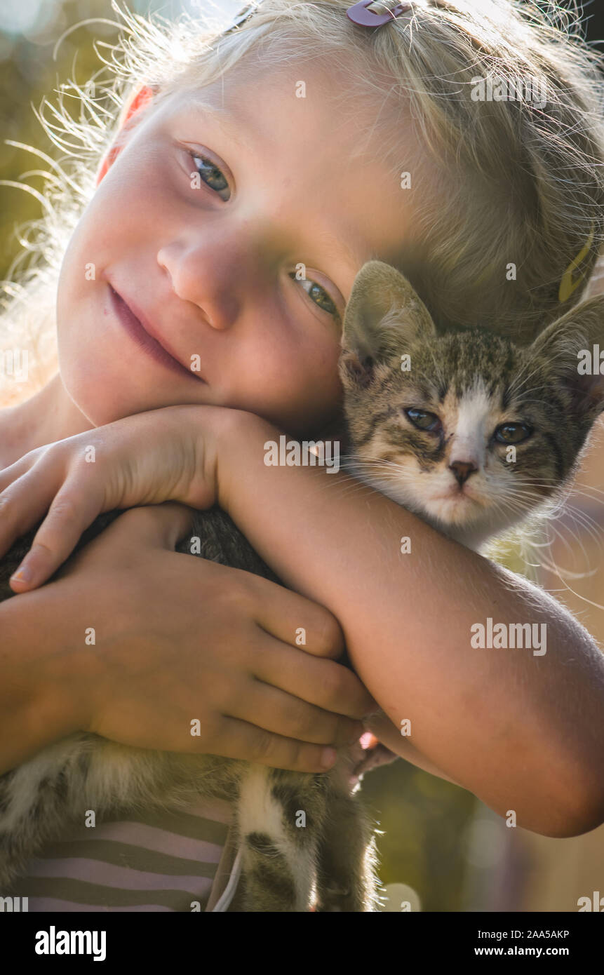 Belle fille blonde holding et caressant petit chat animal assis ensemble dans l'herbe verte sous l'arbre Banque D'Images