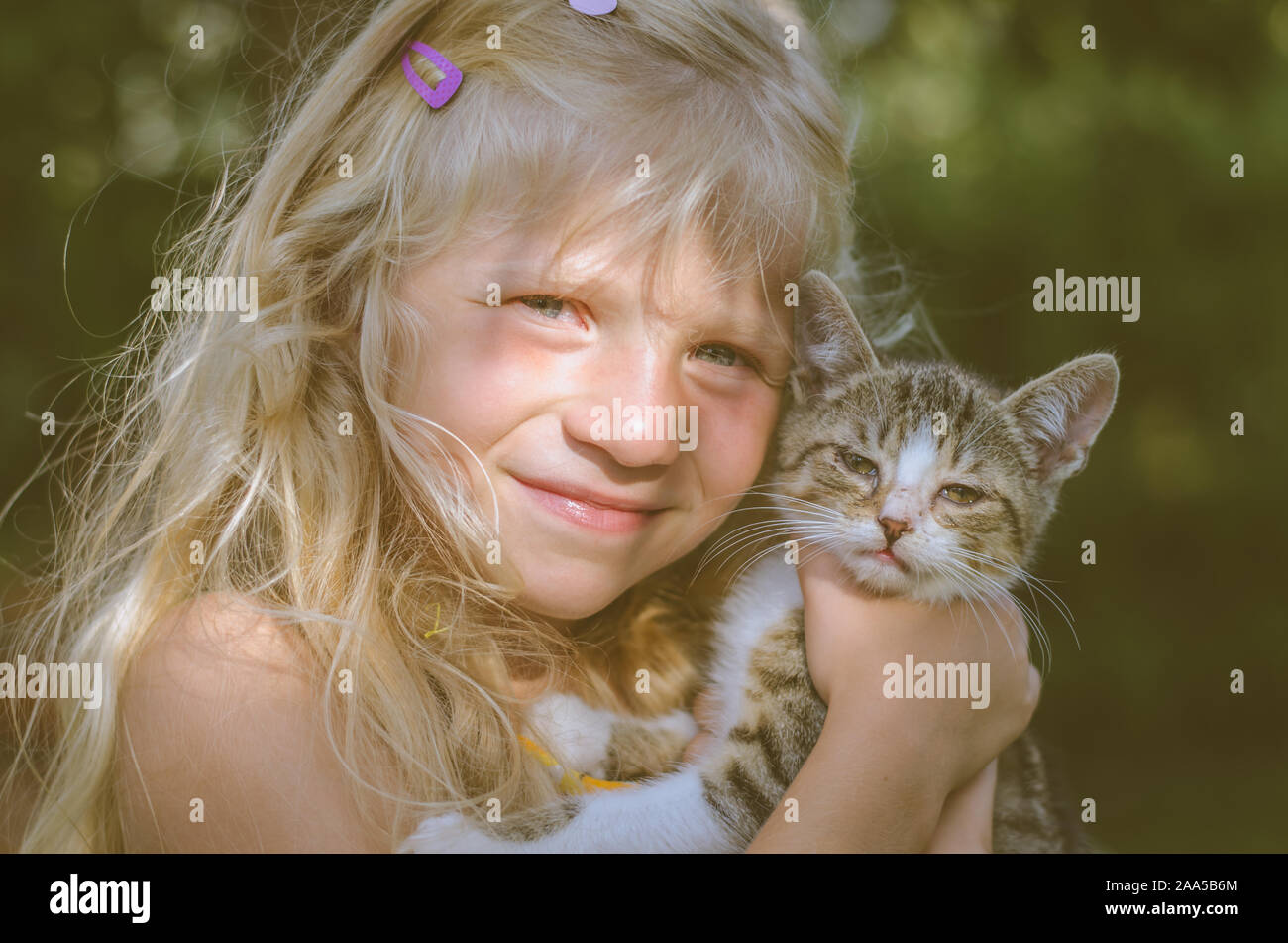 Belle fille blonde holding et caressant petit chat animal assis ensemble dans l'herbe verte sous l'arbre Banque D'Images