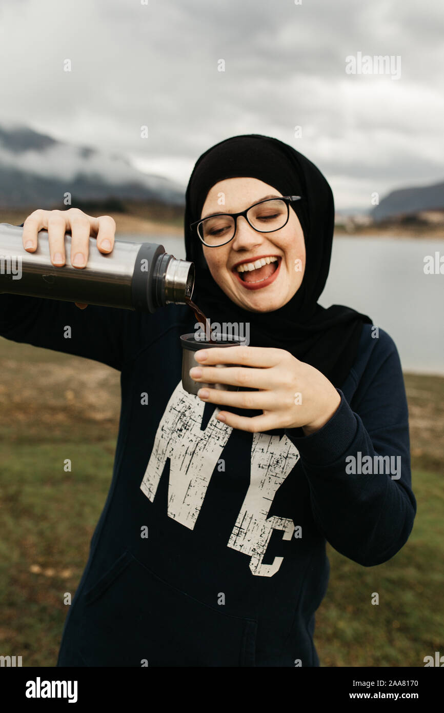 Femme heureuse avec hijab de boire du café dans la nature, sur un matin brumeux lac et montagnes sont à l'arrière-plan. Elle sourit et se plonger le café. Banque D'Images