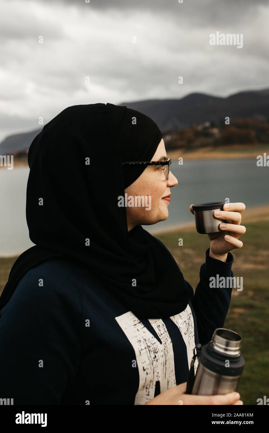 Femme heureuse avec hijab de boire du café dans la nature, sur un matin brumeux, après la pluie. Lac et montagnes sont à l'arrière-plan. Banque D'Images