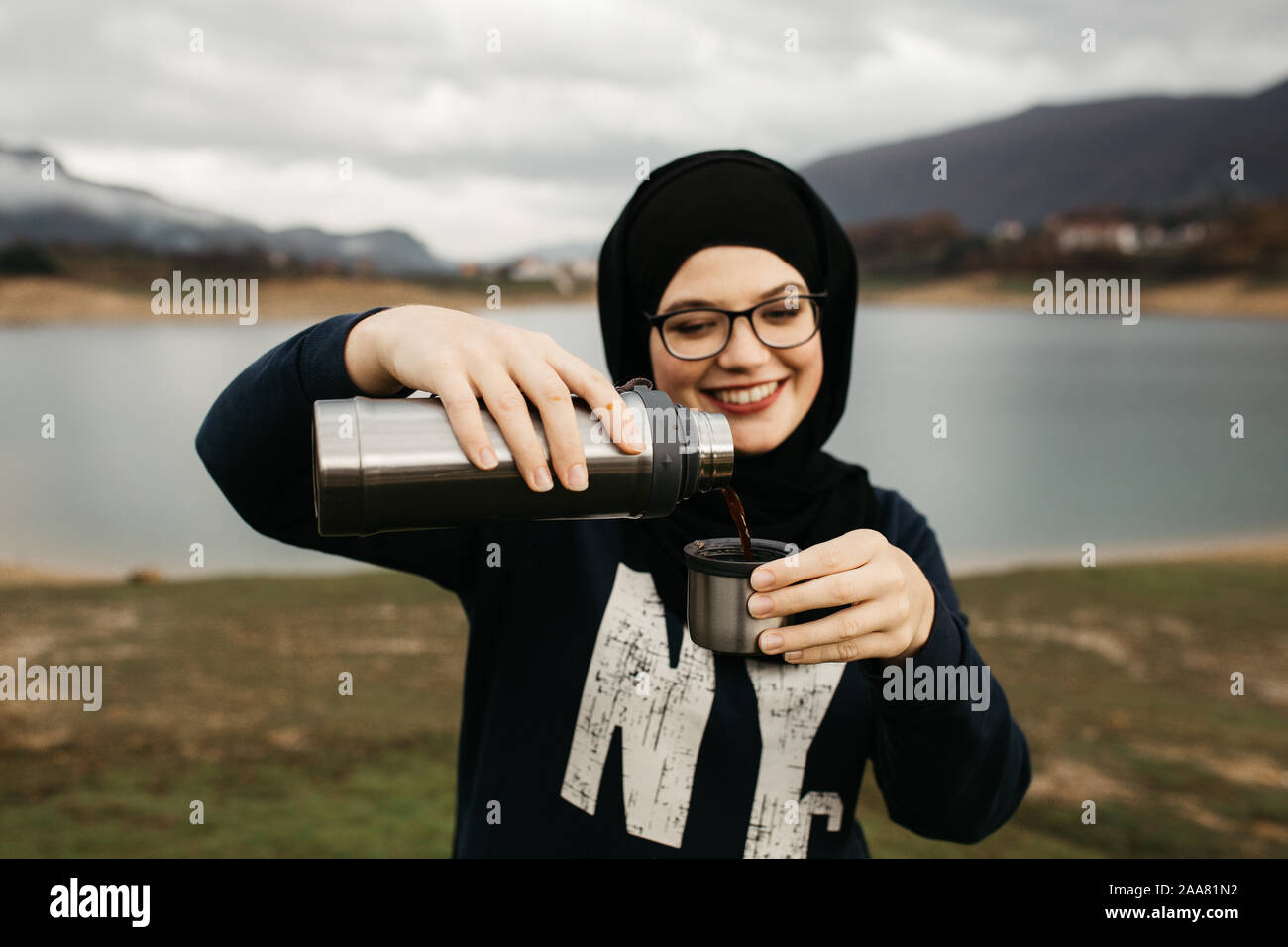 Femme heureuse avec hijab de boire du café dans la nature, sur un matin brumeux lac et montagnes sont à l'arrière-plan. Elle sourit et se plonger le café. Banque D'Images