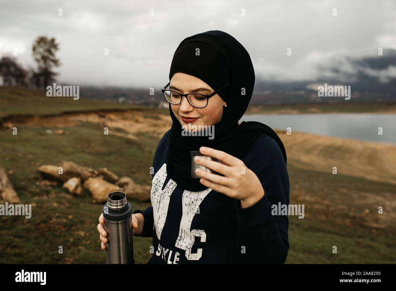 Femme heureuse avec hijab de boire du café dans la nature, sur un matin brumeux, après la pluie. Lac et montagnes sont à l'arrière-plan. Banque D'Images