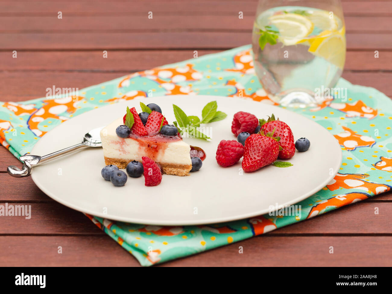 Cheesecake aux fruits rouges, coulis et menthe et verre de l'eau glacée et de citron sur table en bois avec une serviette verte Banque D'Images