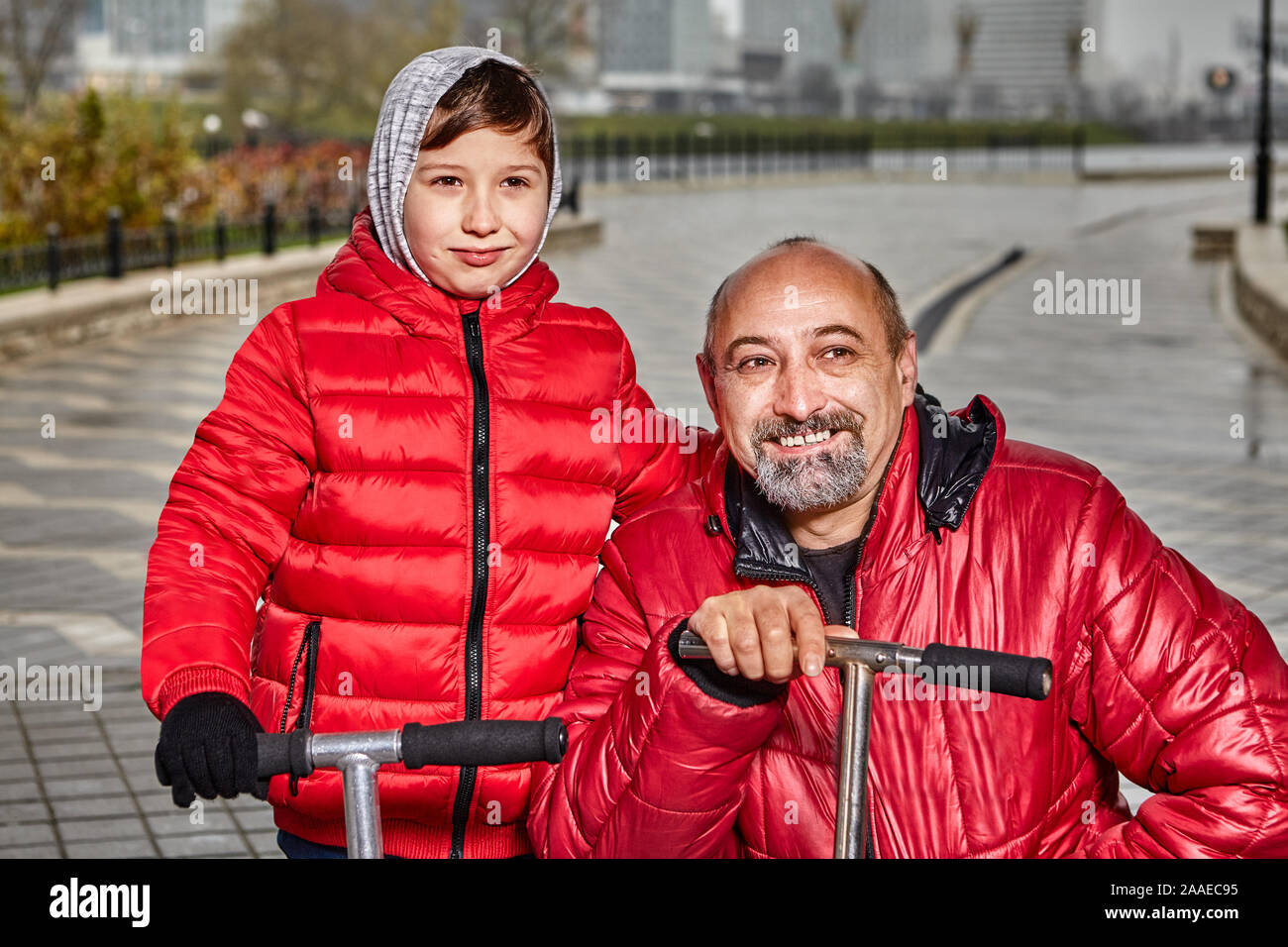 Père et fils ride scooters par temps froid, un garçon de 7 ans et l'homme d'âge moyen vêtus de vêtements chauds à pied le long d'une rue de la ville sous la pluie. Une fam Banque D'Images