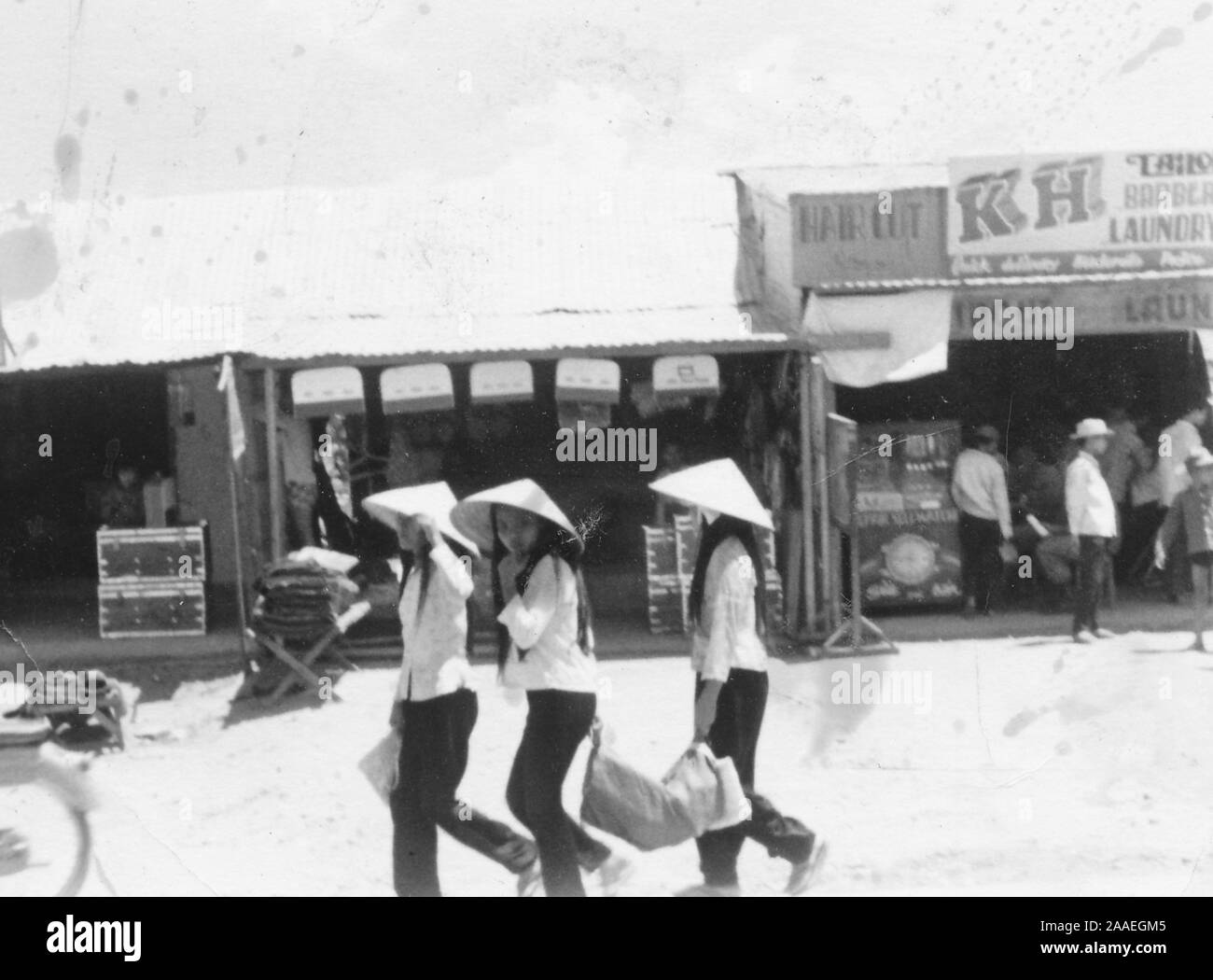 Trois jeunes filles, portant des chemises, des pantalons, et non conique la chapeaux, marcher ensemble à travers une rue commerçante la réalisation d'un objet lourd, avec la fille du milieu en tournant son visage avec curiosité vers l'appareil photo, un China, Vietnam, 1966. () Banque D'Images