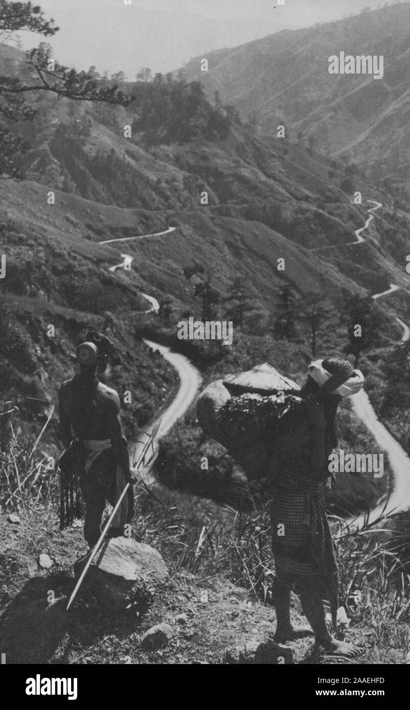 Carte postale monochrome de deux agriculteurs, l'un portant dans un panier de paille sur le dos, dans une zone montagneuse, la Mountain Province, l'île de Luzon, aux Philippines, en 1915. À partir de la Bibliothèque publique de New York. () Banque D'Images