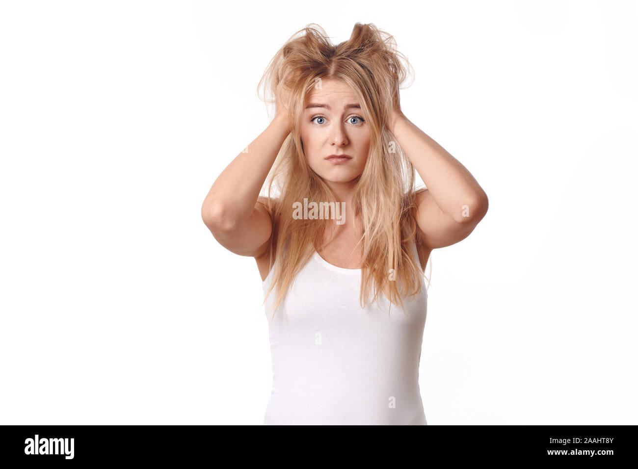 Jeune femme en chemise blanche déçu avec ses cheveux blonds ébouriffés et malpropre, debout avec ses bras à sa tête, looking at camera with sad emoti Banque D'Images