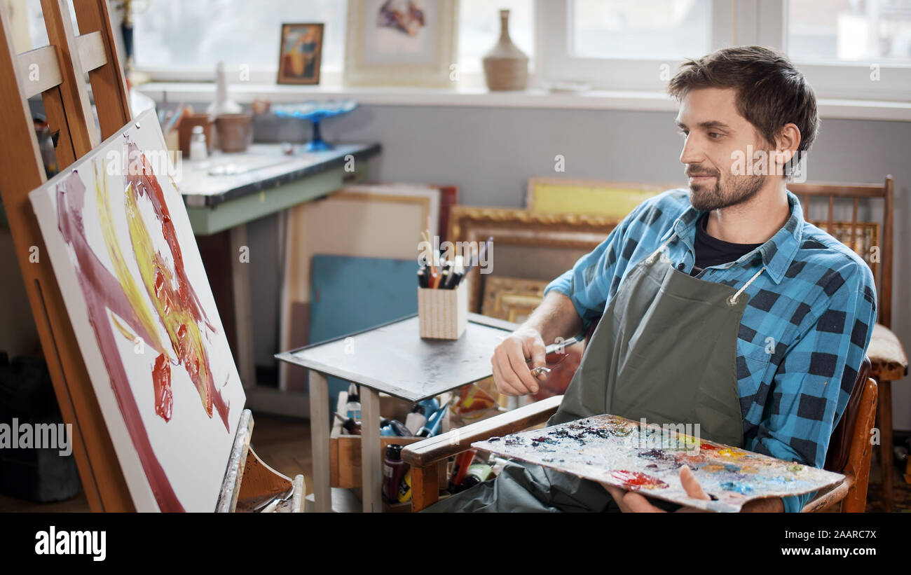 Artiste souriant calme avec une palette et un couteau à palette l'estimation des toiles, il travaille à Banque D'Images