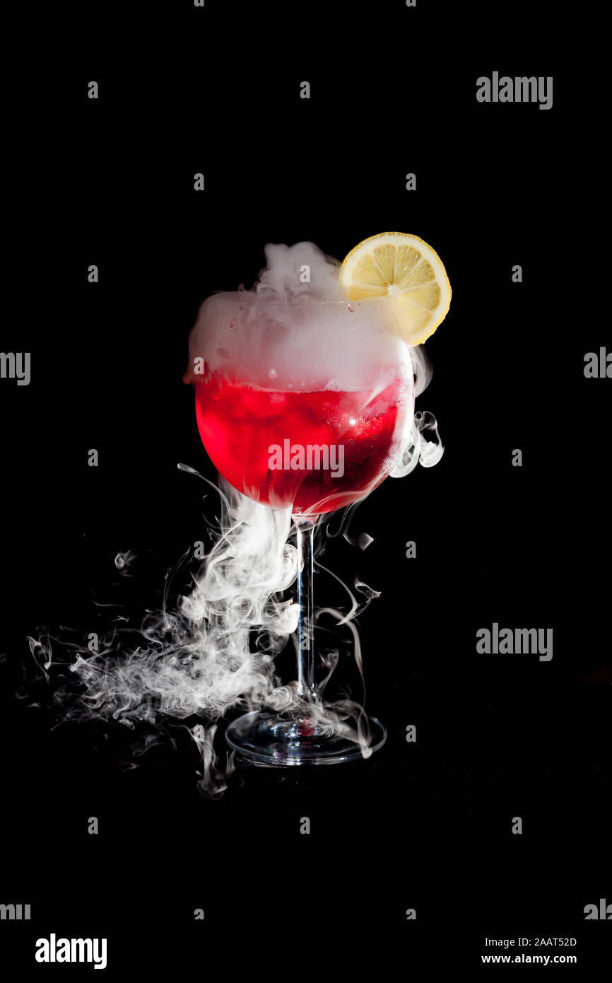Une tasse de cocktail froid rouge avec de la glace sèche fumée et une tranche de citron et fond noir Banque D'Images