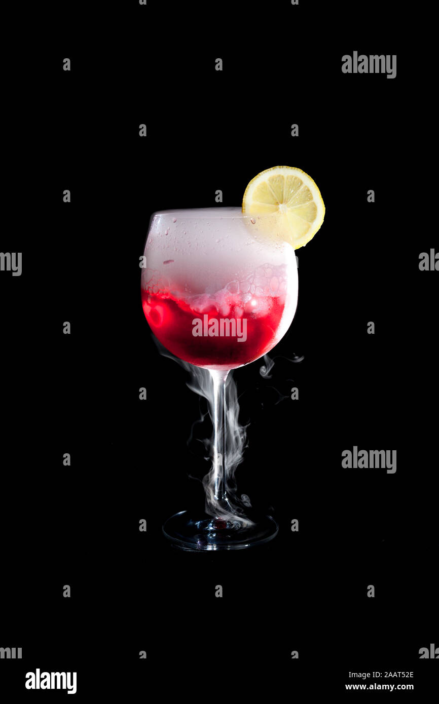 Une tasse de cocktail froid rouge avec de la glace sèche fumée et une tranche de citron et fond noir Banque D'Images