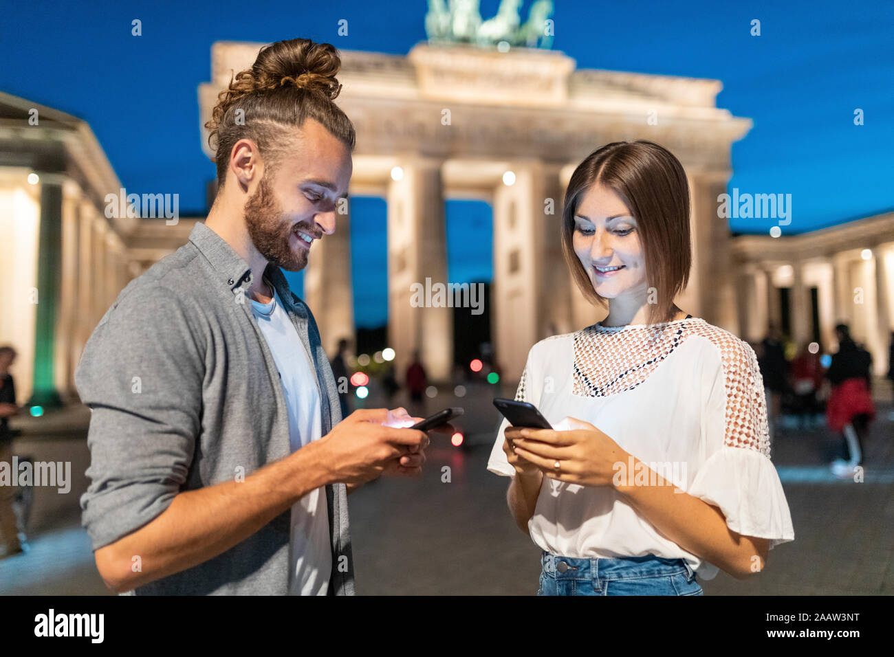 Heureux couple using smartphones à porte de Brandebourg à l'heure bleue, Berlin, Allemagne Banque D'Images