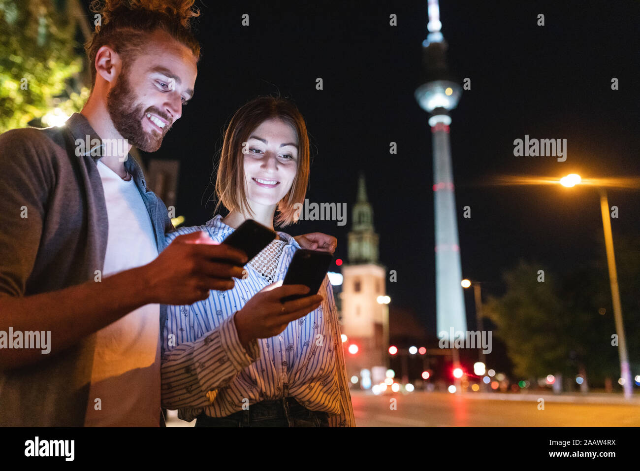 Couple heureux à l'aide de smartphones dans la ville la nuit, Fernsehturm en arrière-plan, Berlin, Allemagne Banque D'Images