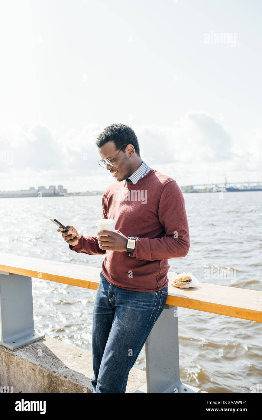 Jeune homme debout à la mer, holding coffee, using smartphone Banque D'Images
