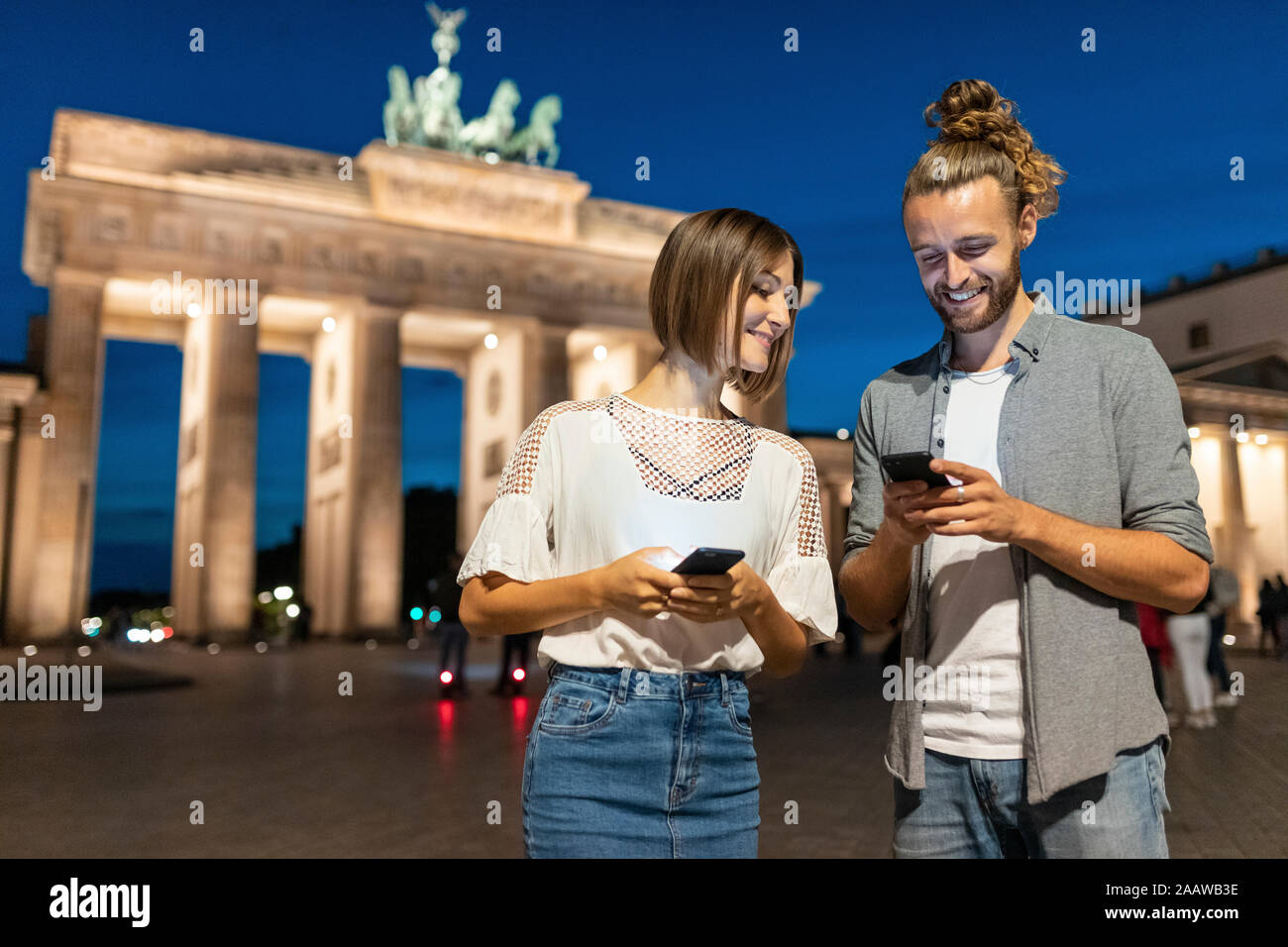 Heureux couple using smartphones à porte de Brandebourg la nuit, Berlin, Allemagne Banque D'Images