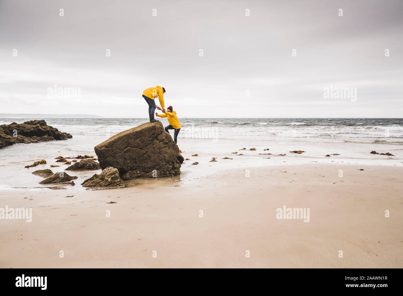 Jeune femme portant des vestes de pluie jaune et de l'escalade sur un rocher sur la plage, Bretagne, France Banque D'Images
