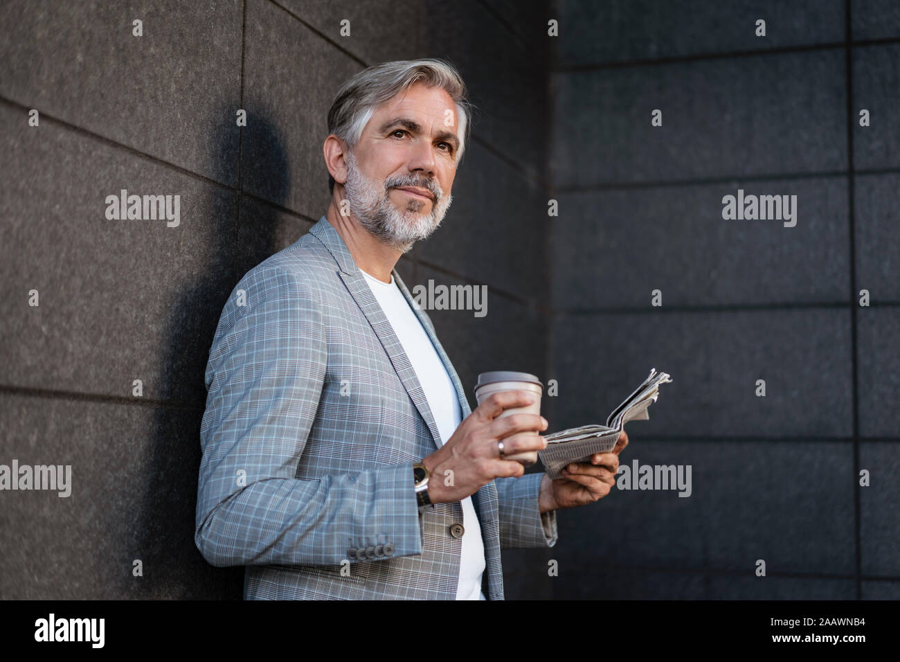 Mature businessman leaning à la mode à un mur avec du papier journal et café à emporter Banque D'Images