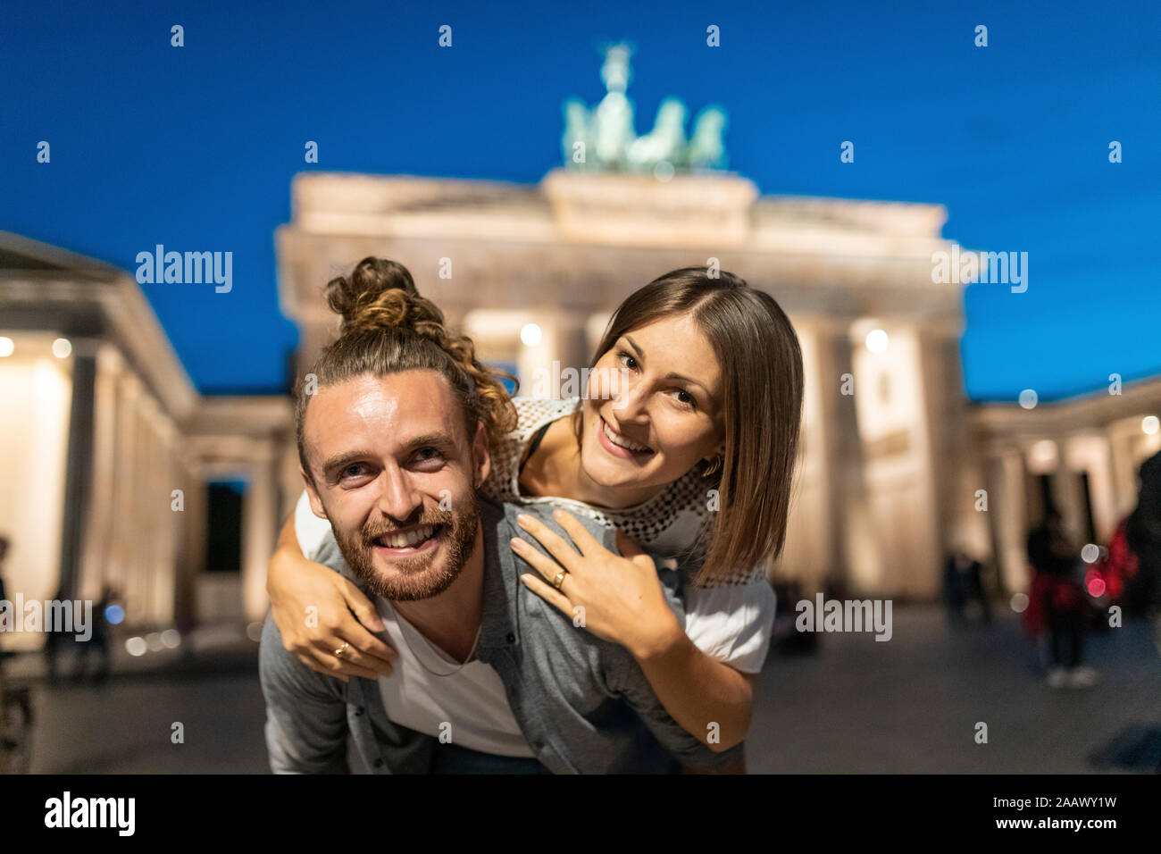 Couple heureux à la porte de Brandebourg à l'heure bleue, jeune homme portant sa petite amie sur son dos, Berlin, Allemagne Banque D'Images