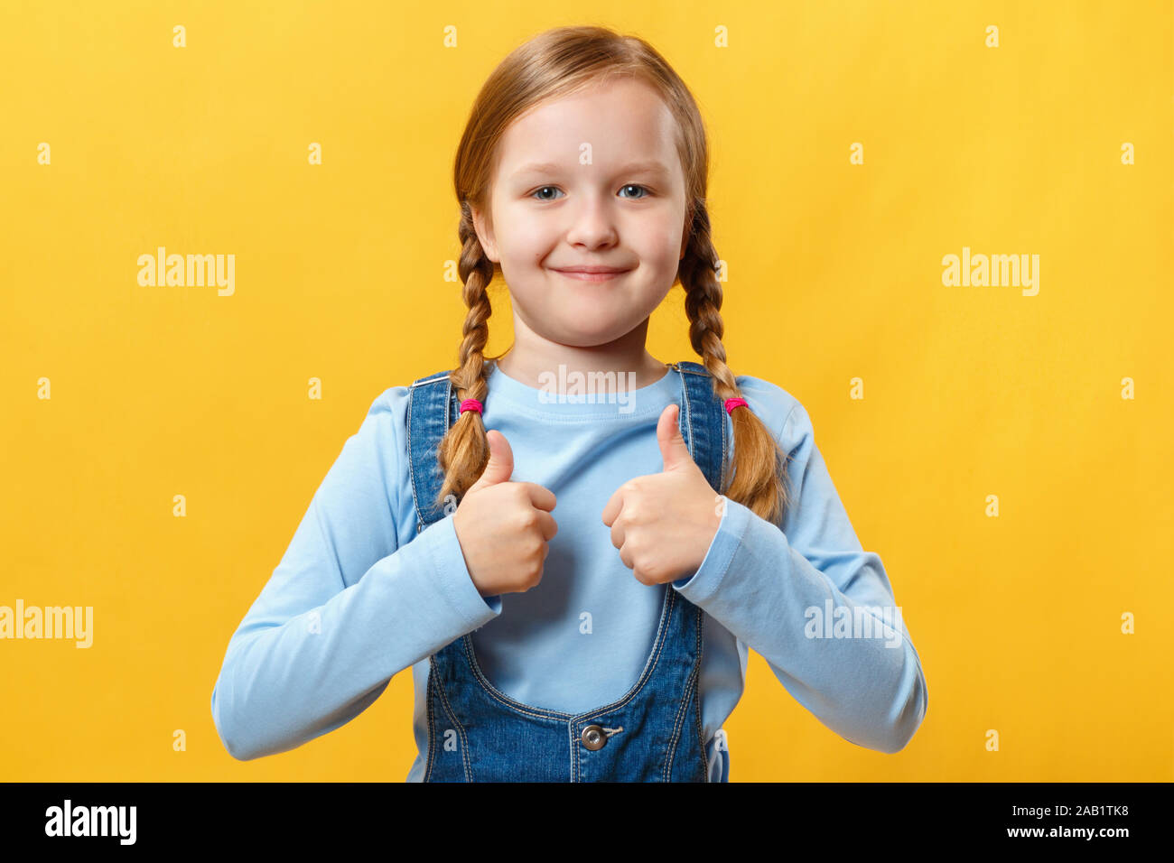 Cute little girl shows thumb up avec deux mains. Un enfant avec des nattes sur fond jaune. Banque D'Images