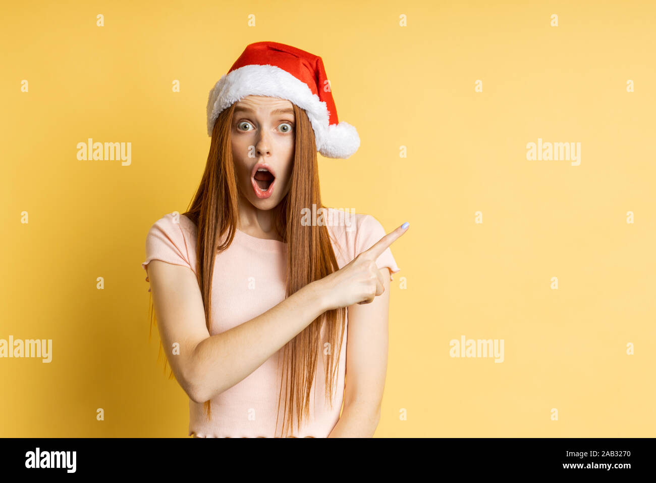 Surpris heureux jeune rousse caucasian woman in Santa hat pointant l'index sur le côté, soulevant les sourcils et ouvrir la bouche, poser sur le mur jaune. C Banque D'Images