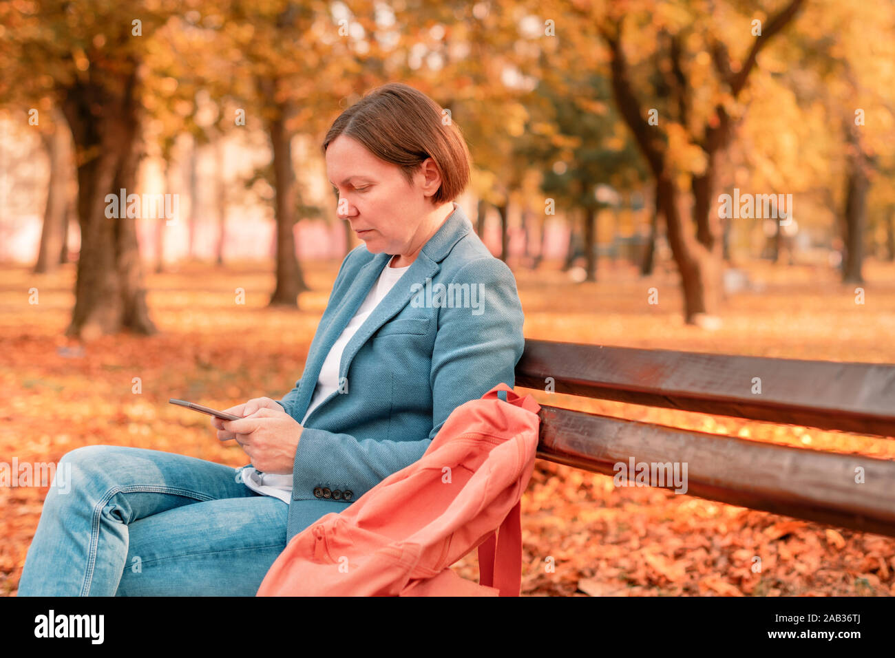 Woman typing text message on mobile phone sur banc de parc d'automne chaud d'automne, aux tons de couleurs image avec selective focus Banque D'Images