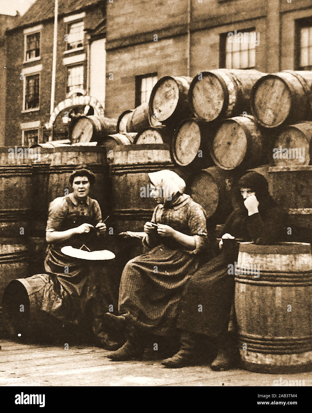 Une vieille photo (début de 1900), de Whitby, North Yorkshire fish femmes en costume traditionnel de détente et des bottes alors que le tricot et le tabagisme chez un tas de barils de harengs sur pier road Banque D'Images