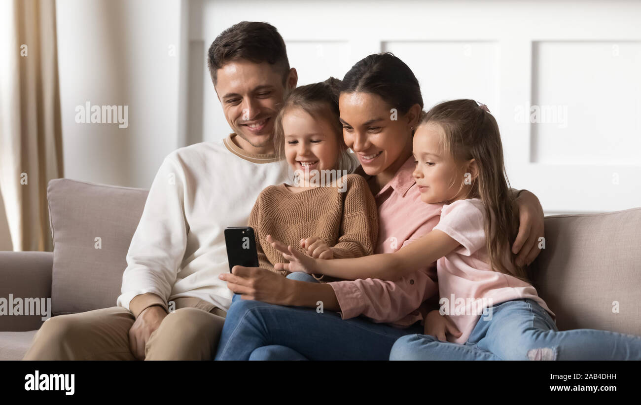 Heureux parents avec enfants filles vous détendre on sofa using smartphone Banque D'Images