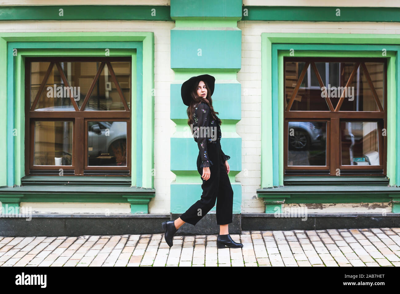 Une jeune fille de race blanche élégante dans des vêtements noirs et un chapeau est la marche dans une ville moderne près de la façade de l'immeuble et regardant la caméra. Banque D'Images