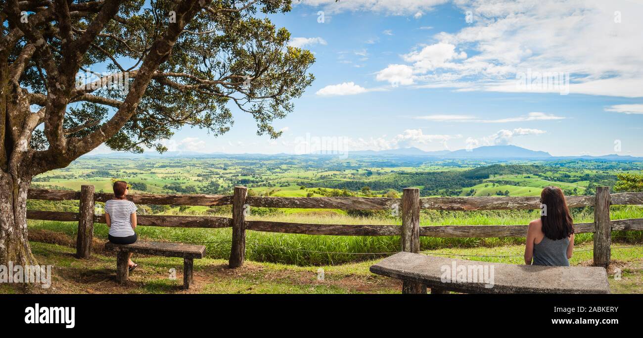 Paysage luxuriant des terres agricoles avec deux femelles assises sur un banc de pierre devant une clôture en bois rustique faite à la main au point de vue de Millaa Millaa, dans le Queensland. Banque D'Images