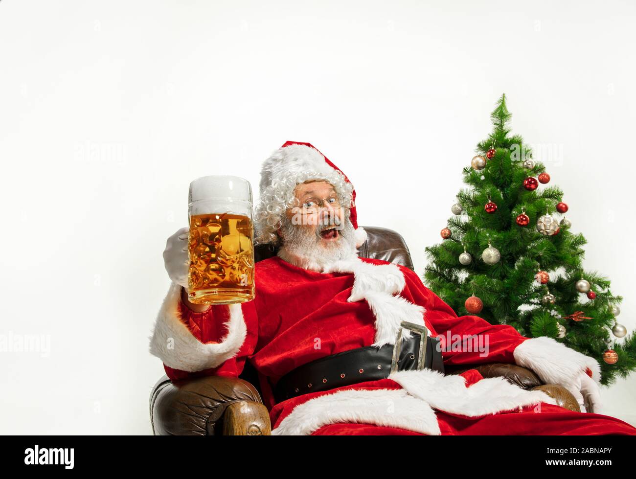 Père Noël bière potable près de l'arbre de Noël, les félicitant, l'air ivre et heureux. Modèle homme de race blanche en costume traditionnel. Nouvelle année 2020, des cadeaux, des vacances, de l'humeur d'hiver. Copyspace pour votre annonce. Banque D'Images