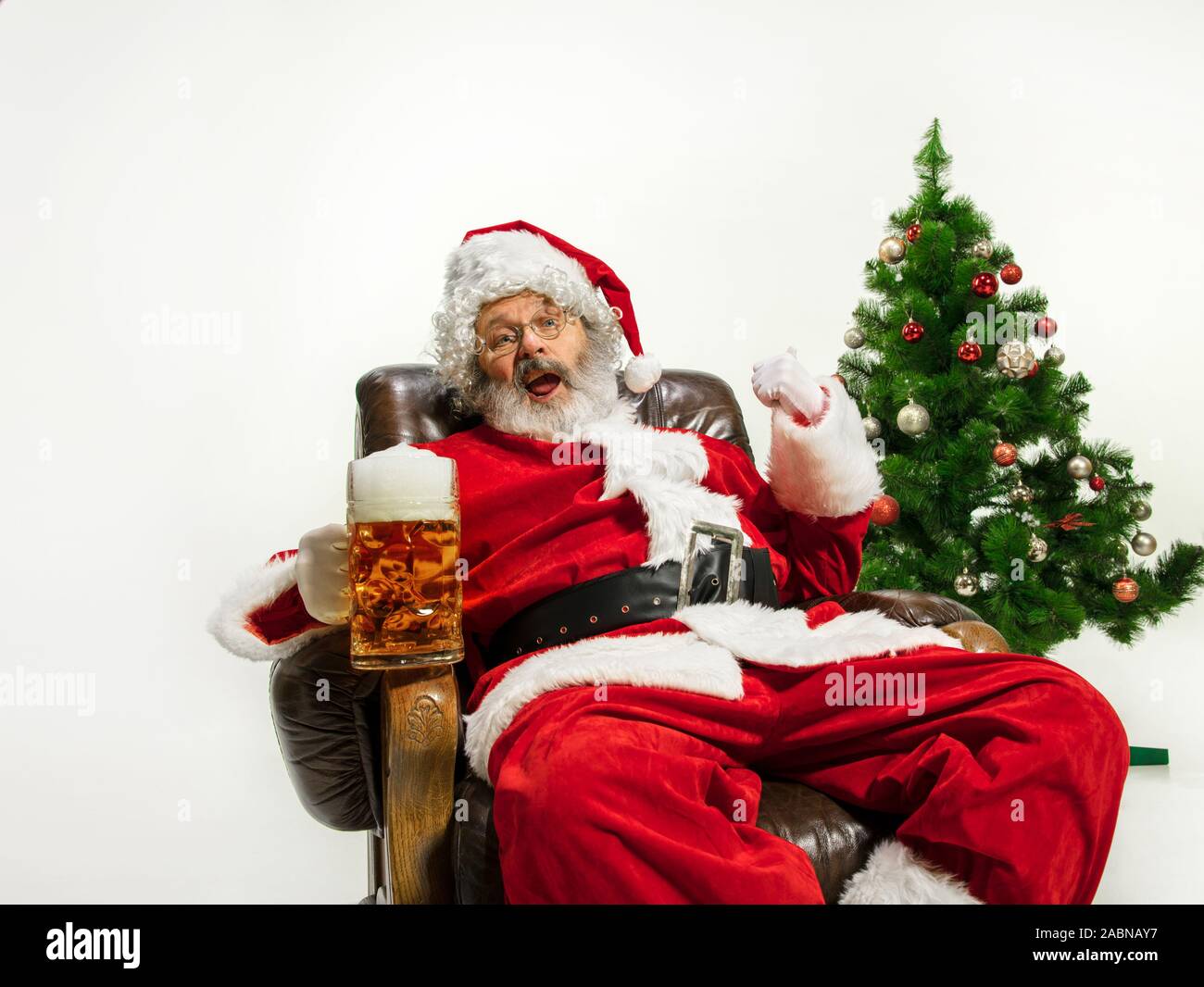 Père Noël bière potable près de l'arbre de Noël, les félicitant, l'air ivre et heureux. Modèle homme de race blanche en costume traditionnel. Nouvelle année 2020, des cadeaux, des vacances, de l'humeur d'hiver. Copyspace pour votre annonce. Banque D'Images