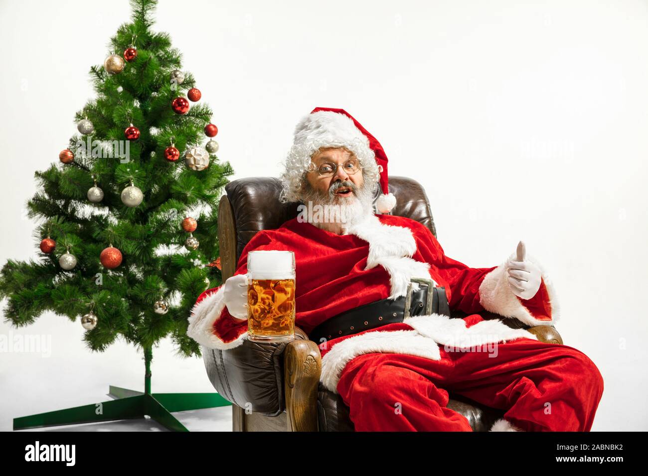 Père Noël bière potable près de l'arbre de Noël, les félicitant, l'air ivre et heureux. Modèle homme de race blanche en costume traditionnel. Nouvelle année 2020, des cadeaux, des vacances, de l'humeur d'hiver. Copyspace pour votre annonce. Banque D'Images