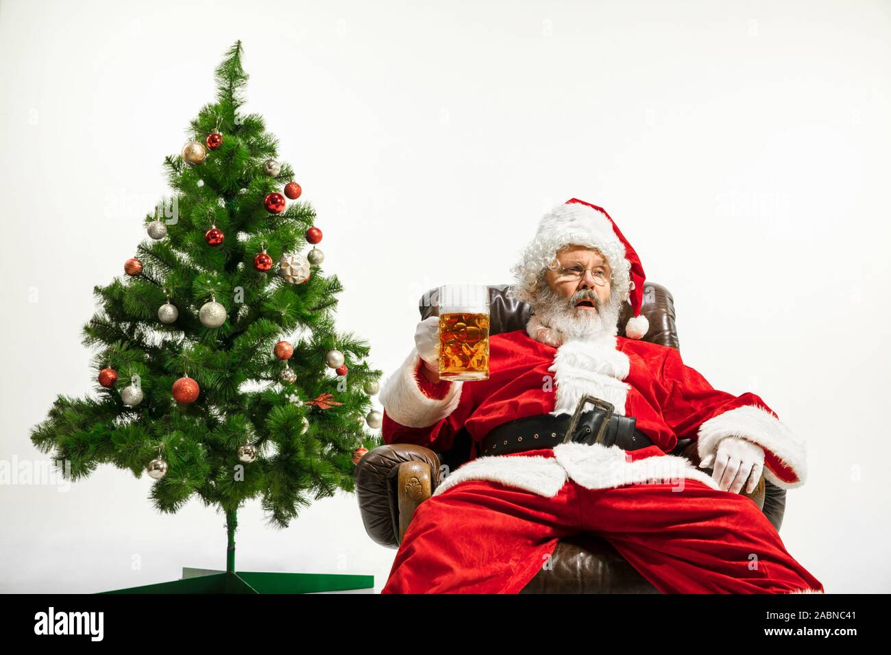 Père Noël bière potable près de l'arbre de Noël, les félicitant, l'air ivre et heureux. Modèle homme de race blanche en costume traditionnel. Nouvelle année 2020, des cadeaux, des vacances, de l'humeur d'hiver. Copyspace pour votre annonce. Banque D'Images