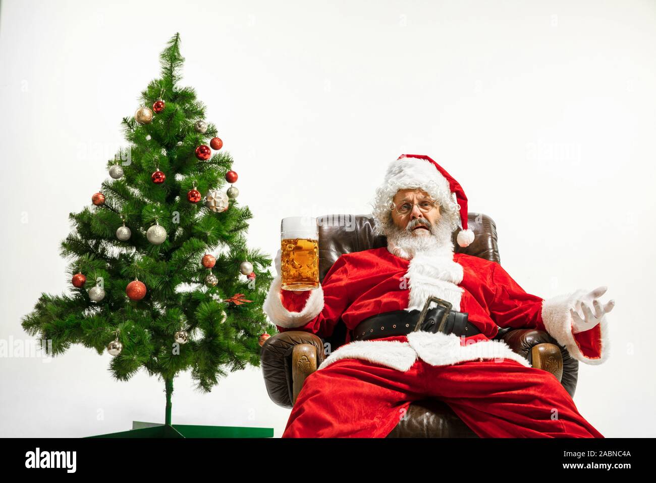 Père Noël bière potable près de l'arbre de Noël, les félicitant, l'air ivre et heureux. Modèle homme de race blanche en costume traditionnel. Nouvelle année 2020, des cadeaux, des vacances, de l'humeur d'hiver. Copyspace pour votre annonce. Banque D'Images