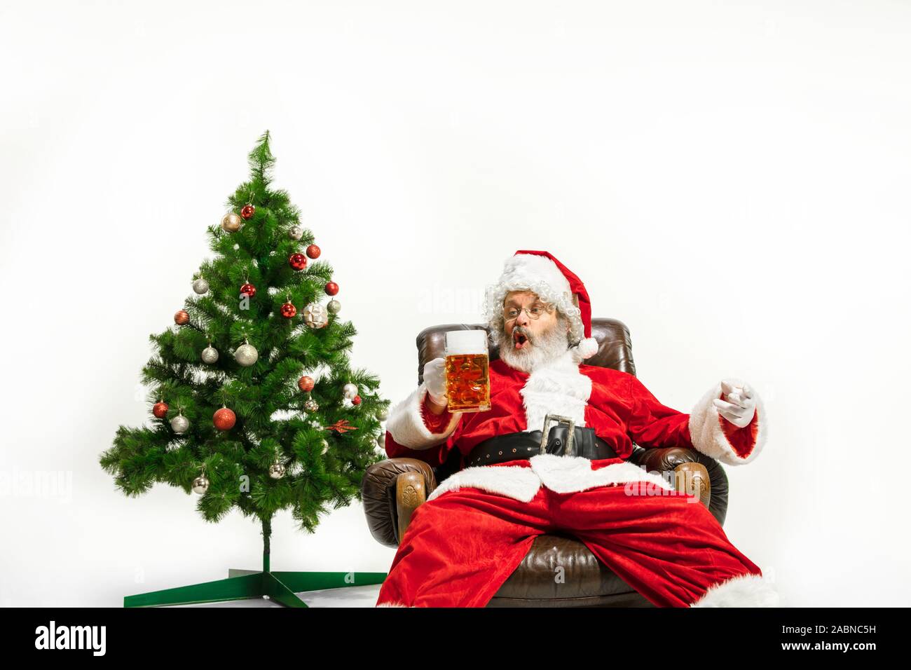 Père Noël bière potable près de l'arbre de Noël, les félicitant, l'air ivre et heureux. Modèle homme de race blanche en costume traditionnel. Nouvelle année 2020, des cadeaux, des vacances, de l'humeur d'hiver. Copyspace pour votre annonce. Banque D'Images