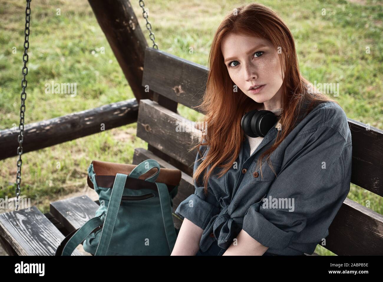 Redhead girl dans une chemise en jean est assis avec un sac à dos sur une balançoire en bois banc et regarde dans le cadre. Banque D'Images