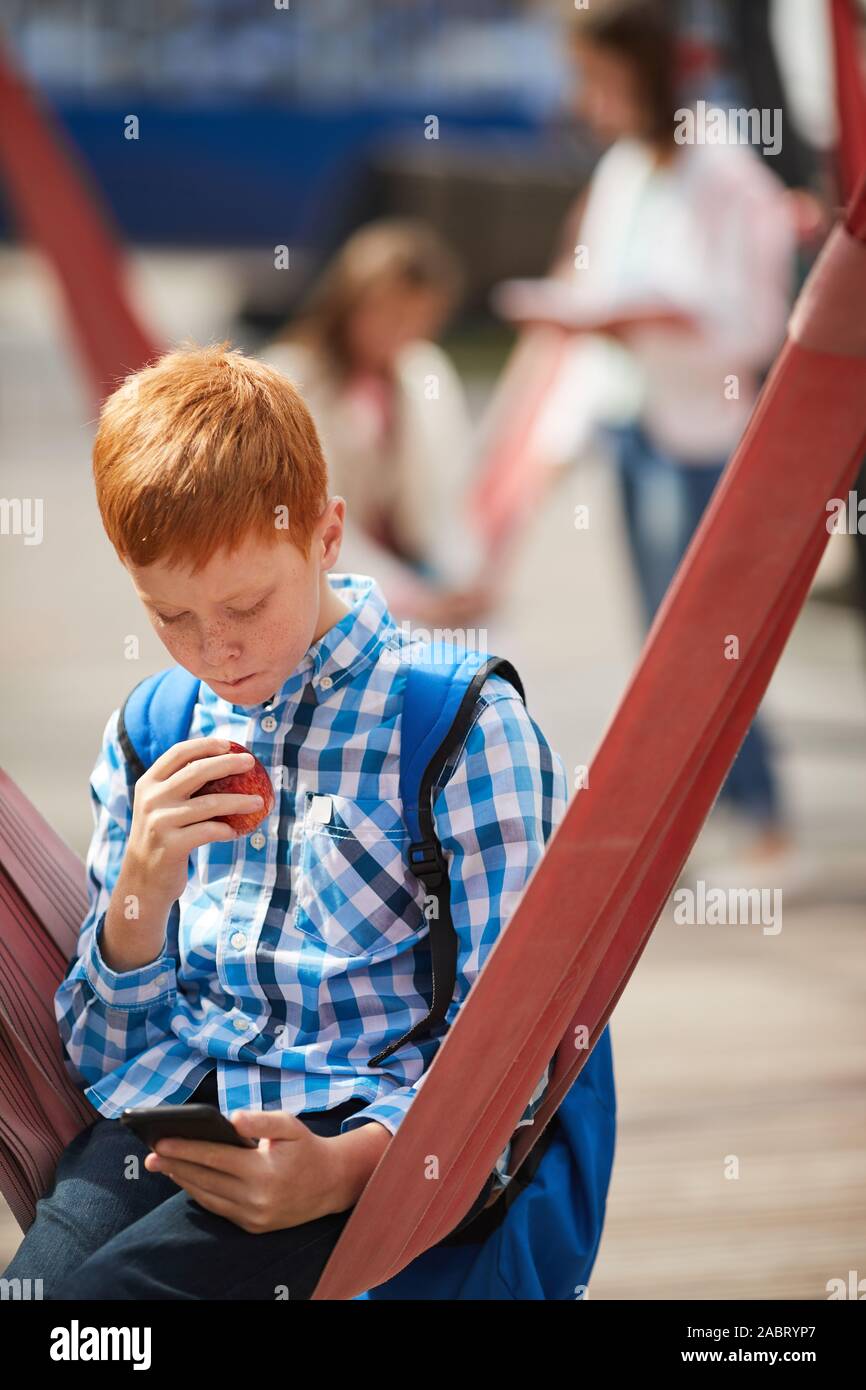 À l'aide d'écolier aux cheveux rouge téléphone mobile et délicieux tout en restant assis sur swing sur le terrain de jeux en plein air Banque D'Images