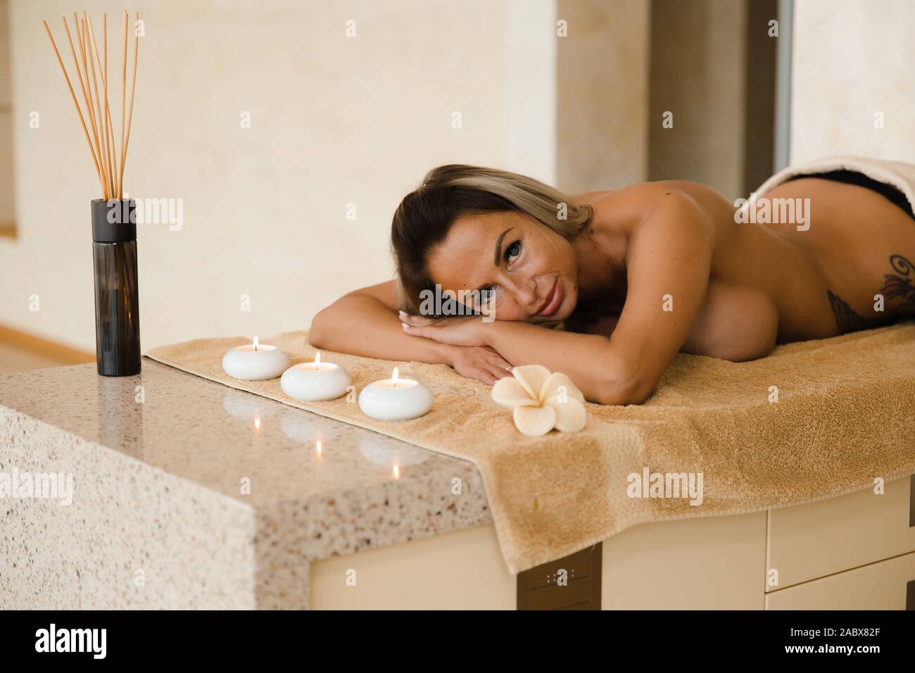 Blonde woman relaxing in salon de couché sur la table de massage avec des bougies et de l'arôme des bâtons. Jeune fille avec des tatouages de vous détendre dans un centre spa concept - Banque D'Images